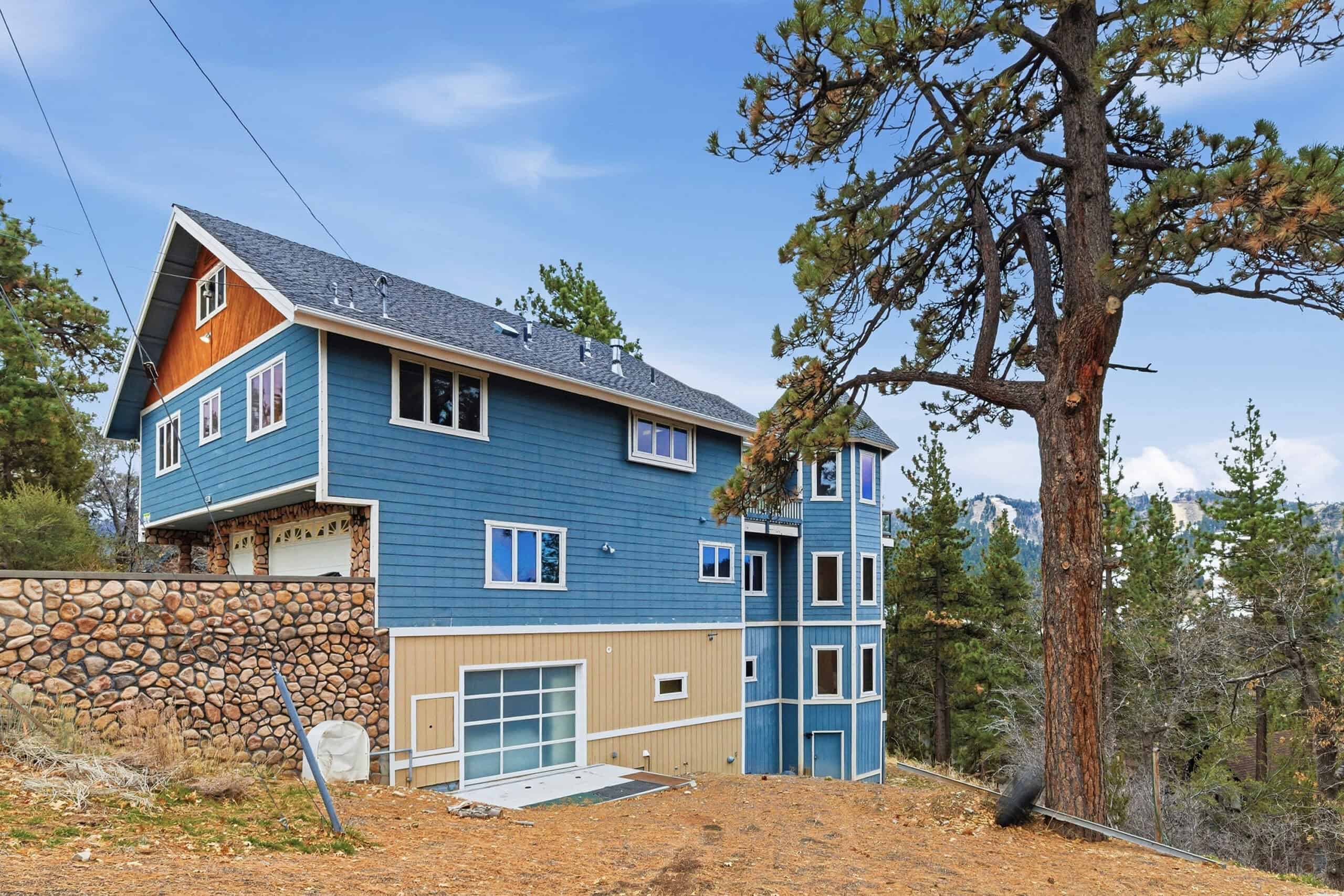 Contemporary blue house with multiple levels and large trees in a wooded area.
