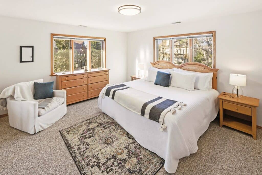 A cozy bedroom with a large bed, wooden furniture, and natural light from two windows.